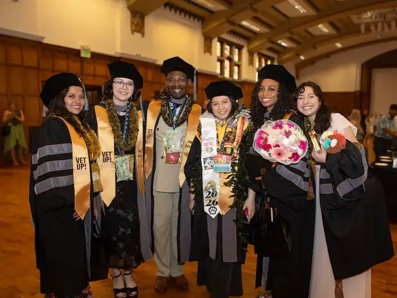 Six graduates in caps and gowns pose together indoors, wearing sashes; two hold bouquets.
